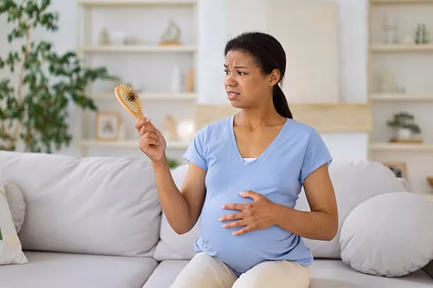 Pregnant woman sitting on a sofa, holding a hairbrush in one hand and resting a hand on her baby bump, looking uncomfortable or concerned.